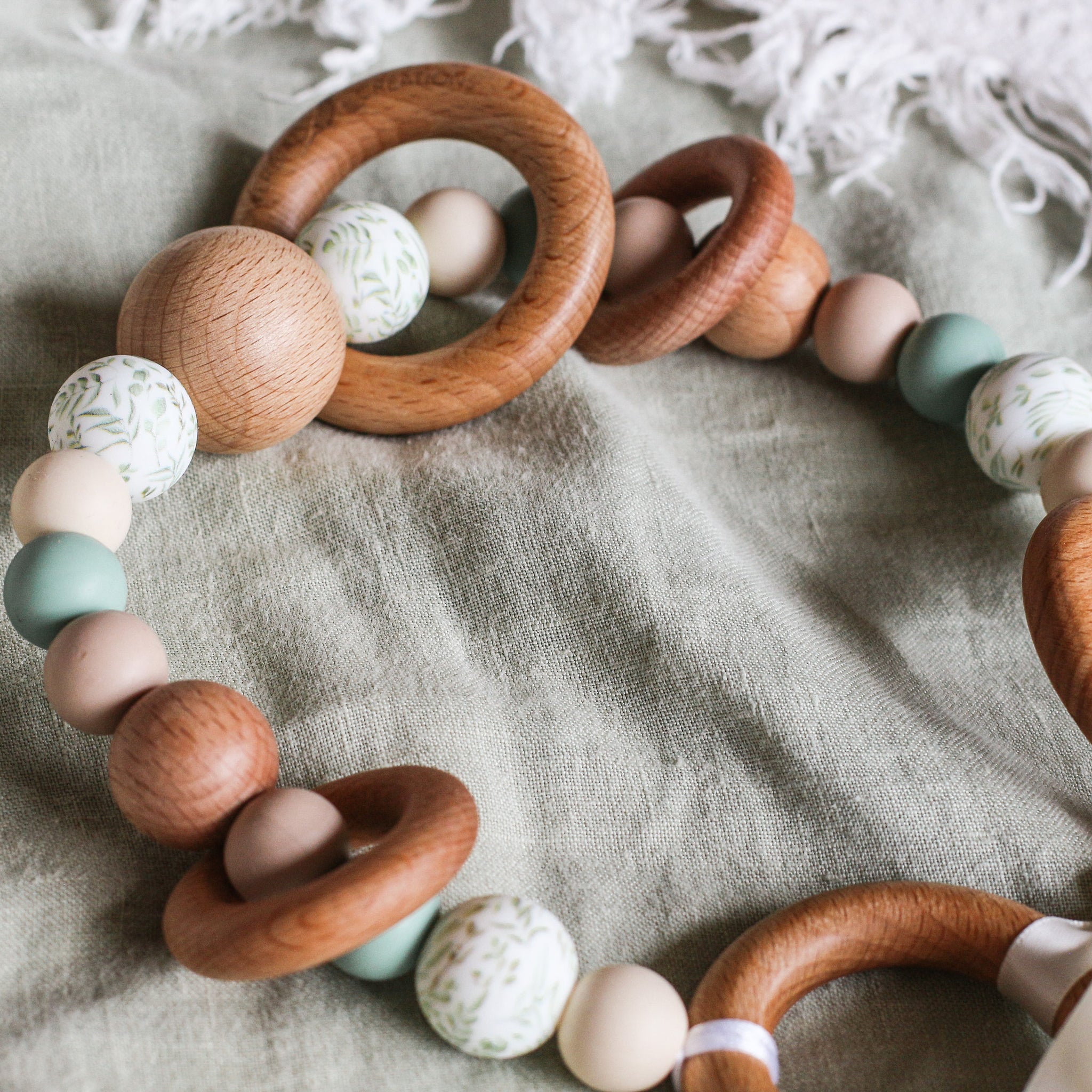 Close up of handmade silicone and wood pram toy in sage, beige and printed leafy beads
