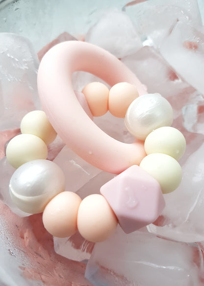 Baby teething toy handmade from silicone sitting in a bowl of ice. Pink, coral, white.