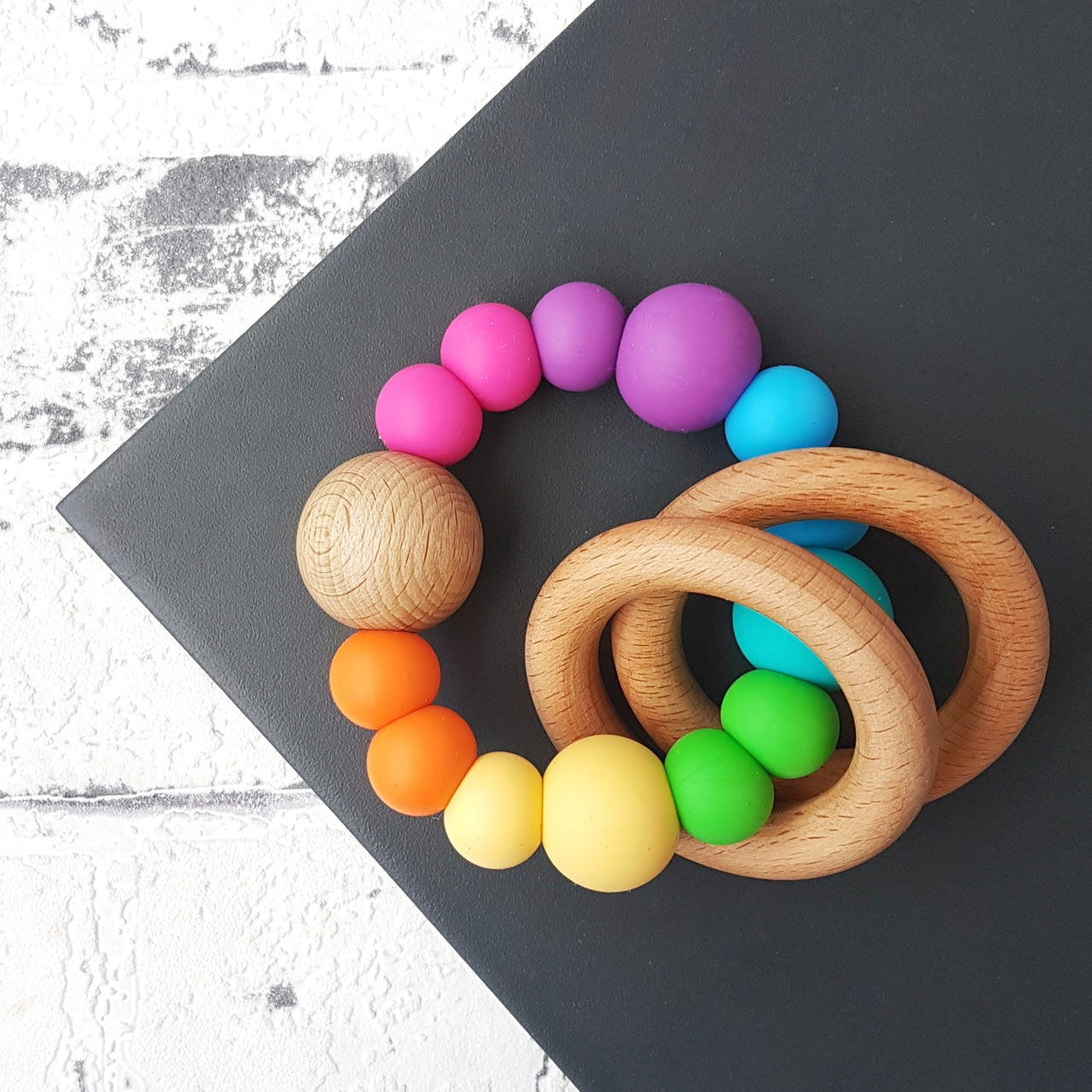 Teether/sensory toy featuring 2 wooden rings that rattle. Handmade from silicone and natural wood. Pastel and muted rainbow. Bright rainbow.